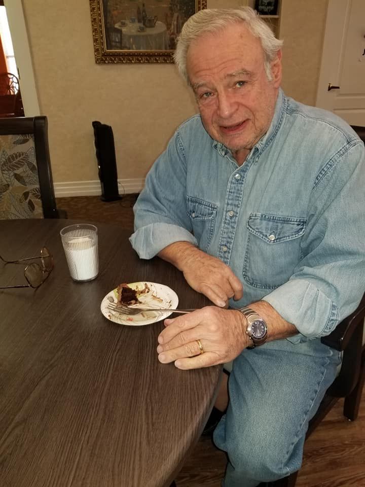 A senior man in a denim shirt sits at a table with a glass of milk and a piece of cake.