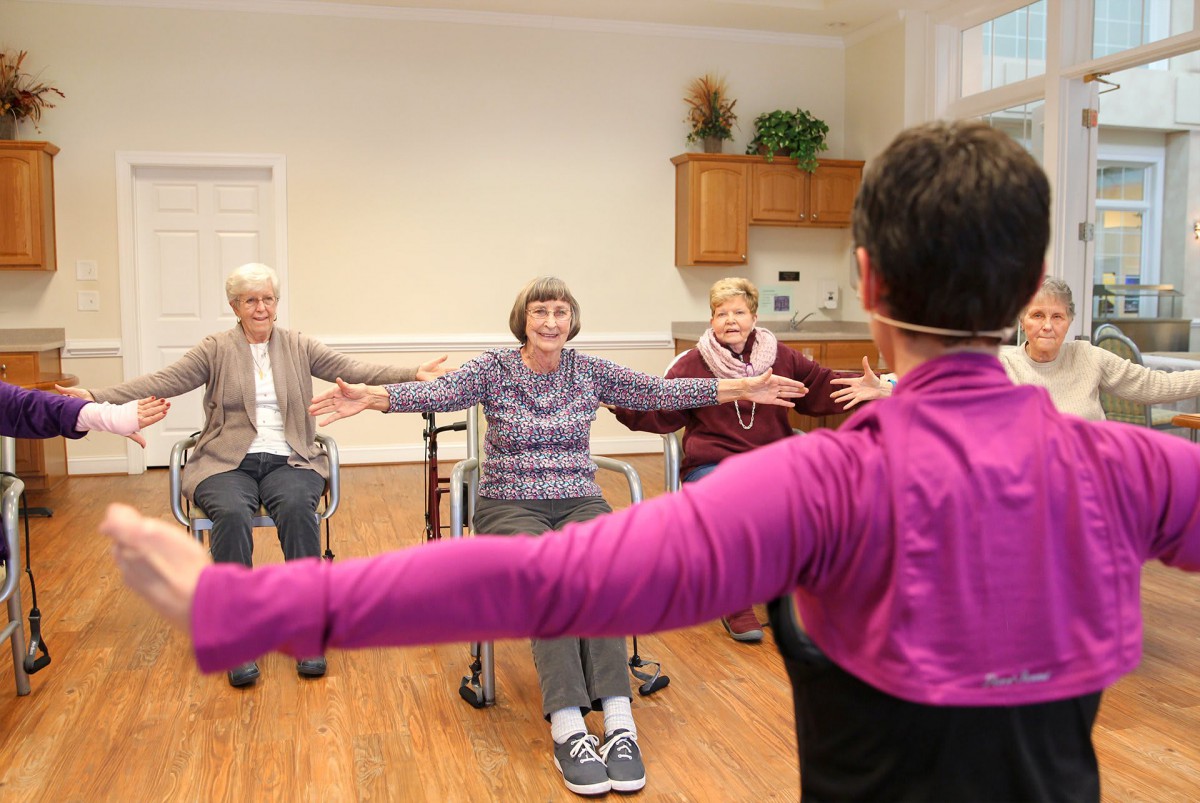 A group of seniors participating in a seated exercise class indoors, led by an instructor.