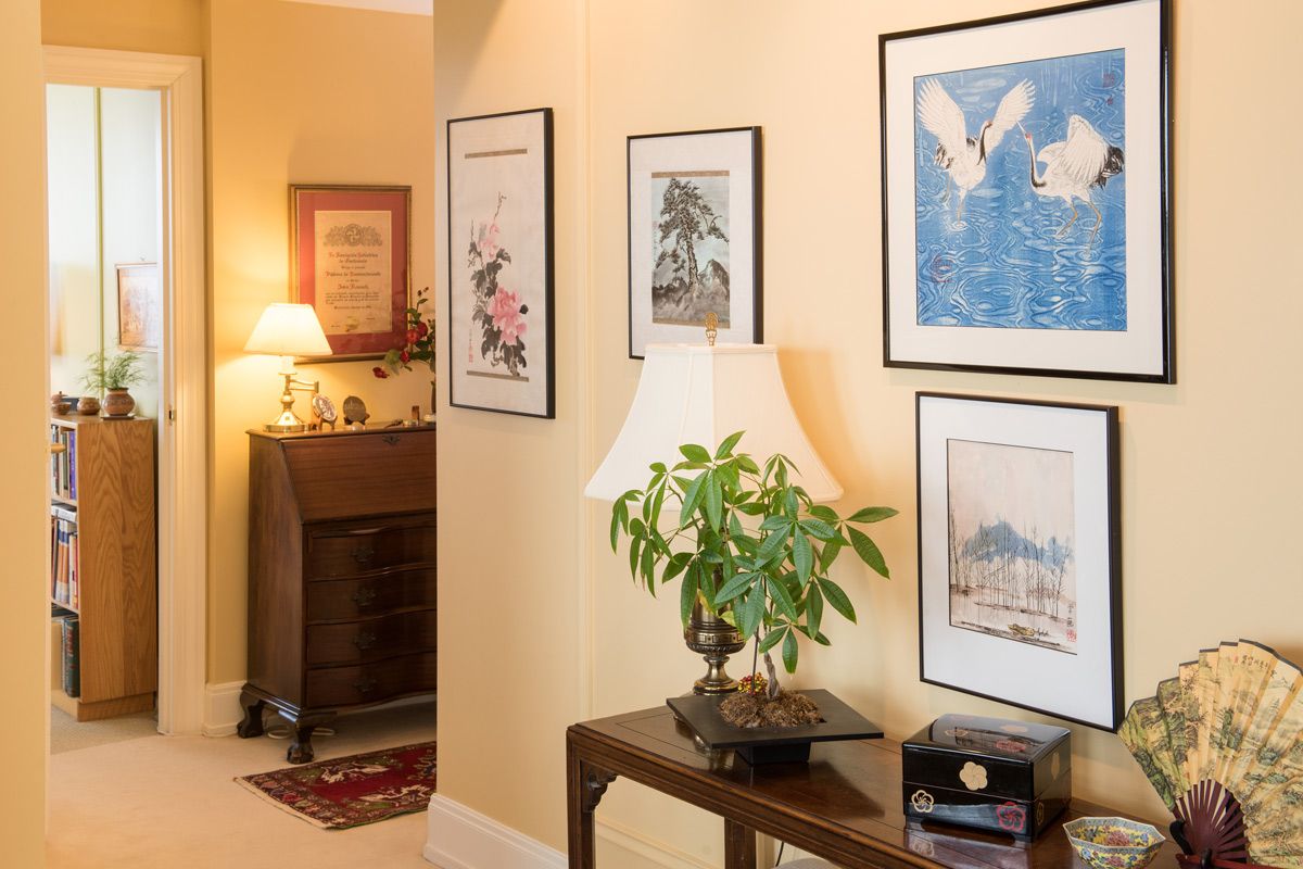 A warmly lit hallway featuring framed artwork, a small table with a lamp and plant, and a cozy corner with furniture in the background.