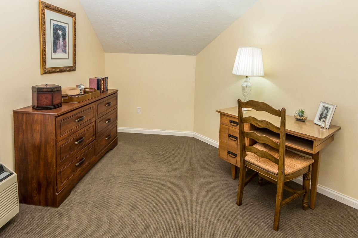 A cozy corner with a desk and chair, accompanied by a dresser and lamp in a carpeted room.