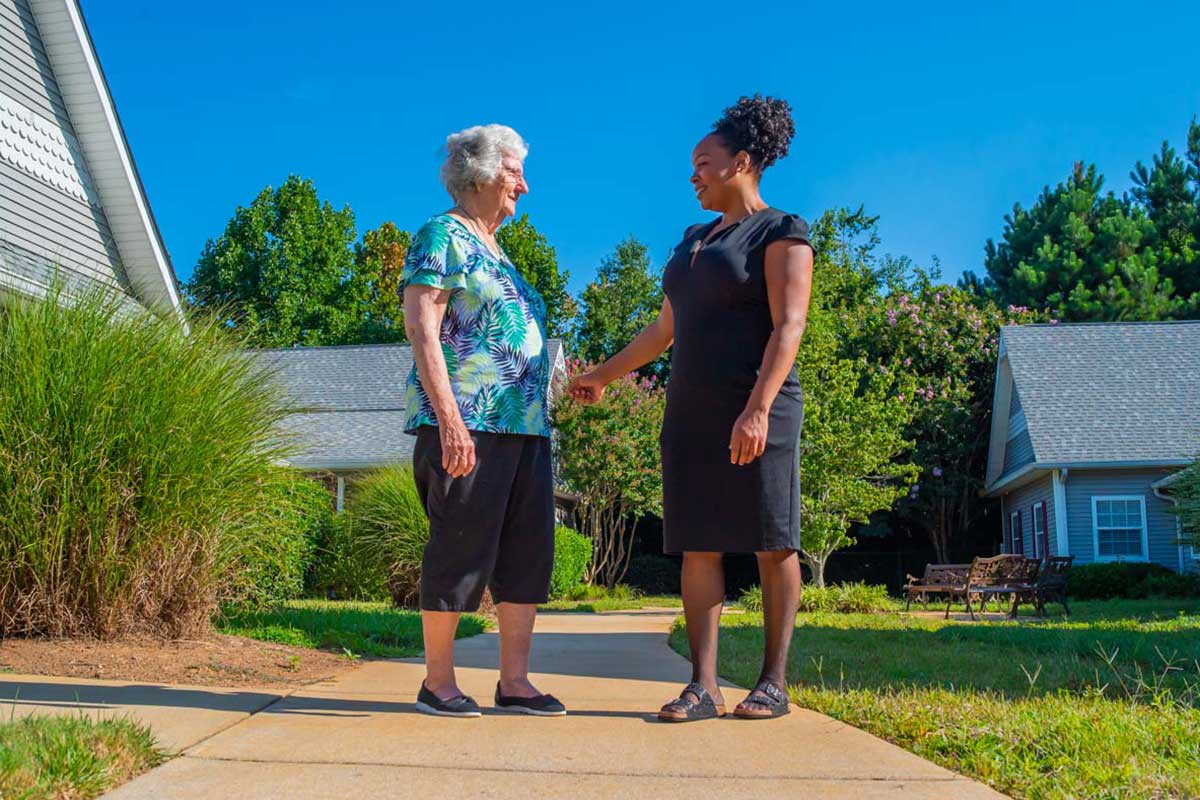 An elderly woman and a younger woman shake hands outdoors on a sunny day.
