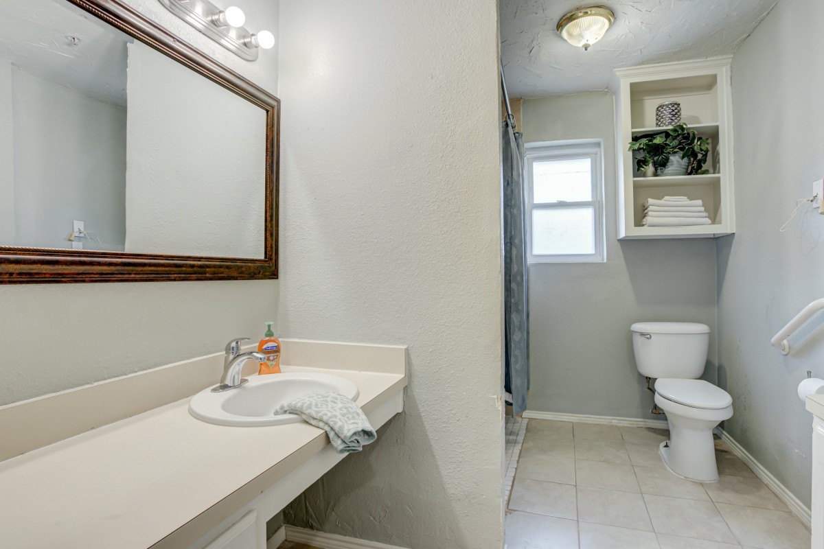 A clean and bright bathroom featuring a sink, mirror, toilet, and window with storage shelves.