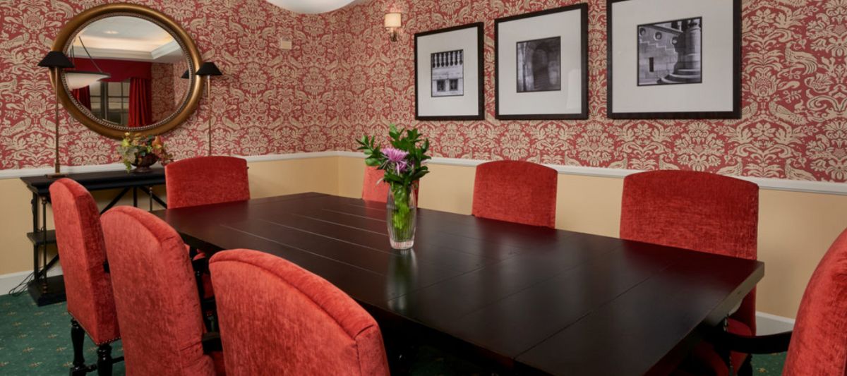 A dining room with red upholstered chairs, a dark wooden table, and a vase of flowers as a centerpiece. The walls are decorated with patterned wallpaper and framed pictures.
