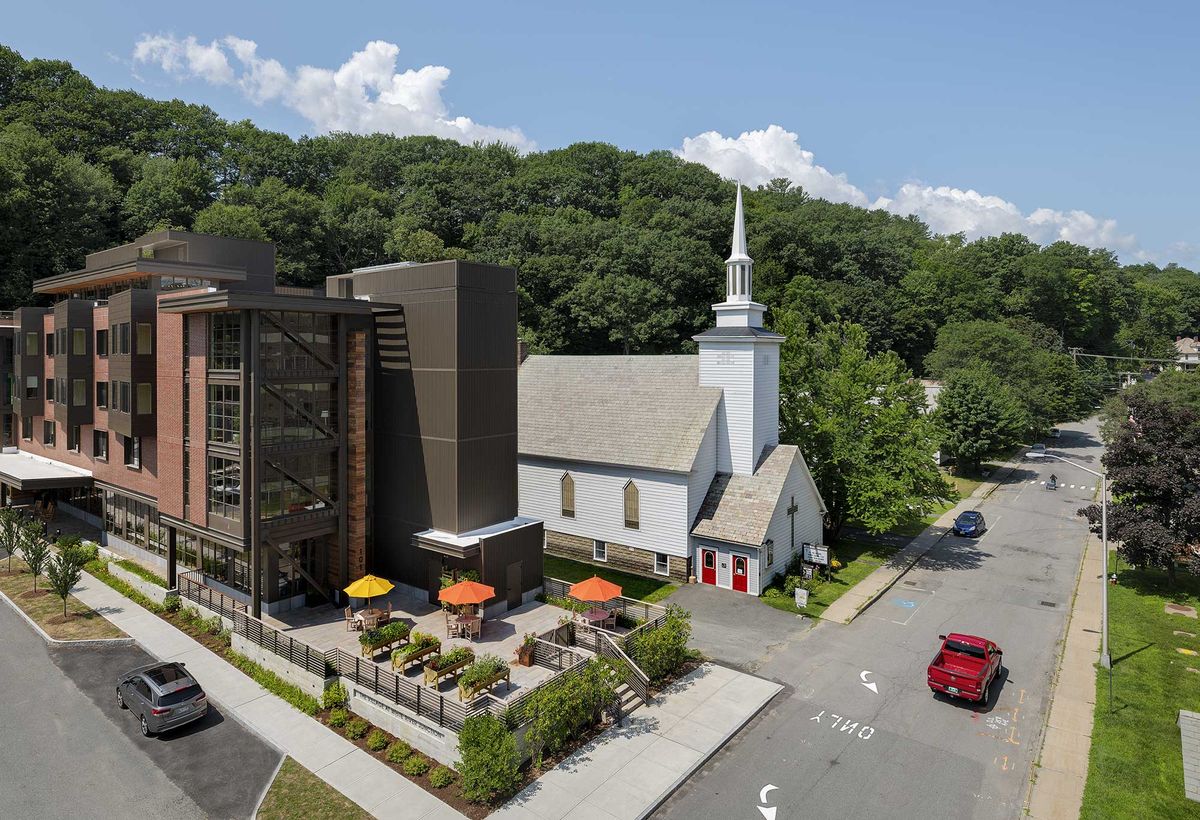 A modern building beside a traditional church with outdoor seating and a parking lot, surrounded by lush trees.