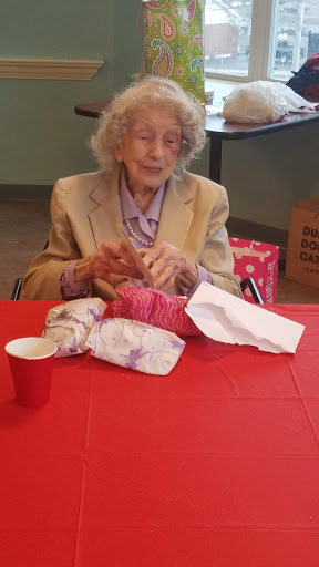 An elderly woman engaged in knitting at a table decorated with a red cloth, surrounded by crafting materials.