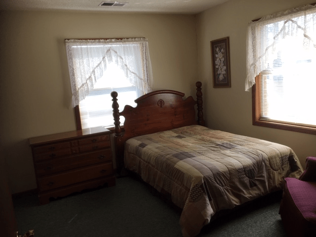 A cozy bedroom with a wooden bed, dresser, and windows adorned with lace curtains.