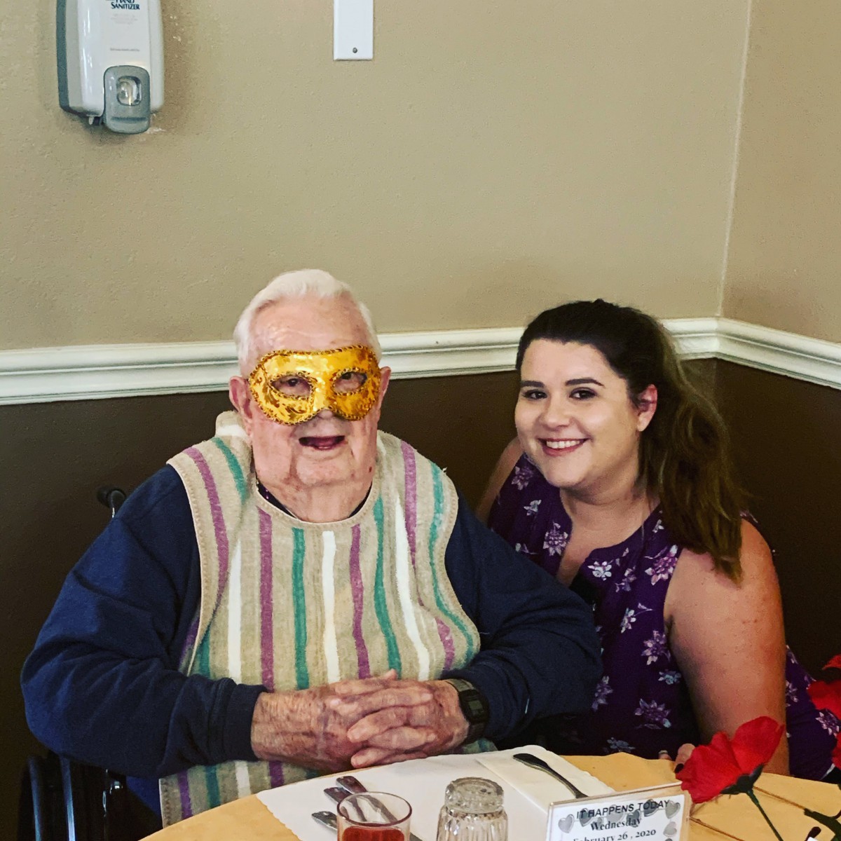 An elderly man wearing a mask and a woman smiling together at a dining table during a celebration.
