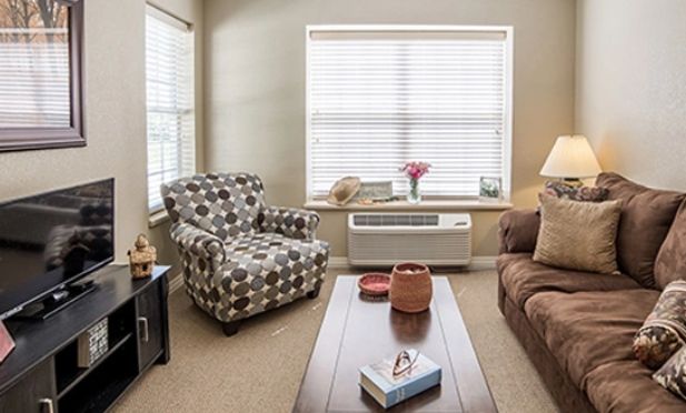 A cozy living room with a patterned armchair, sofa, and coffee table, featuring a large window with blinds.