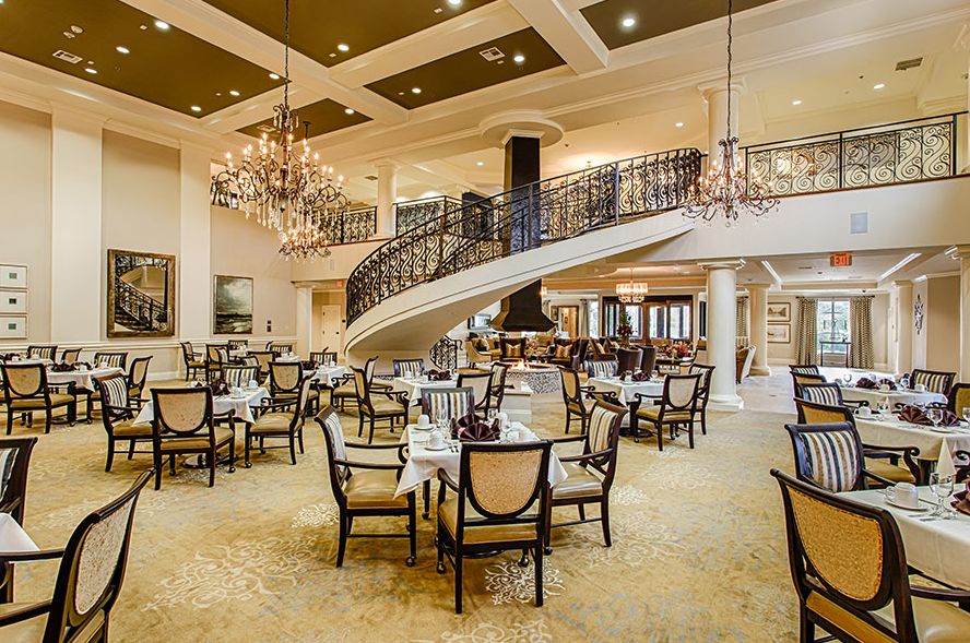 An elegant restaurant interior featuring chandeliers, a grand staircase, and neatly arranged tables and chairs.