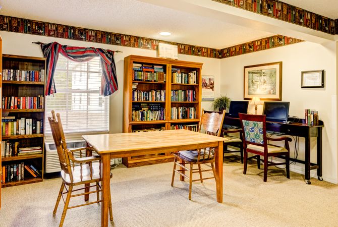A cozy library room featuring a table and chairs, bookshelves filled with books, and a couple of computers by the window.