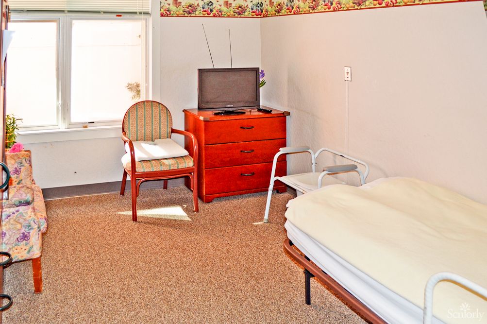 A simple bedroom with basic furniture including a bed, dresser, chairs, and a TV.