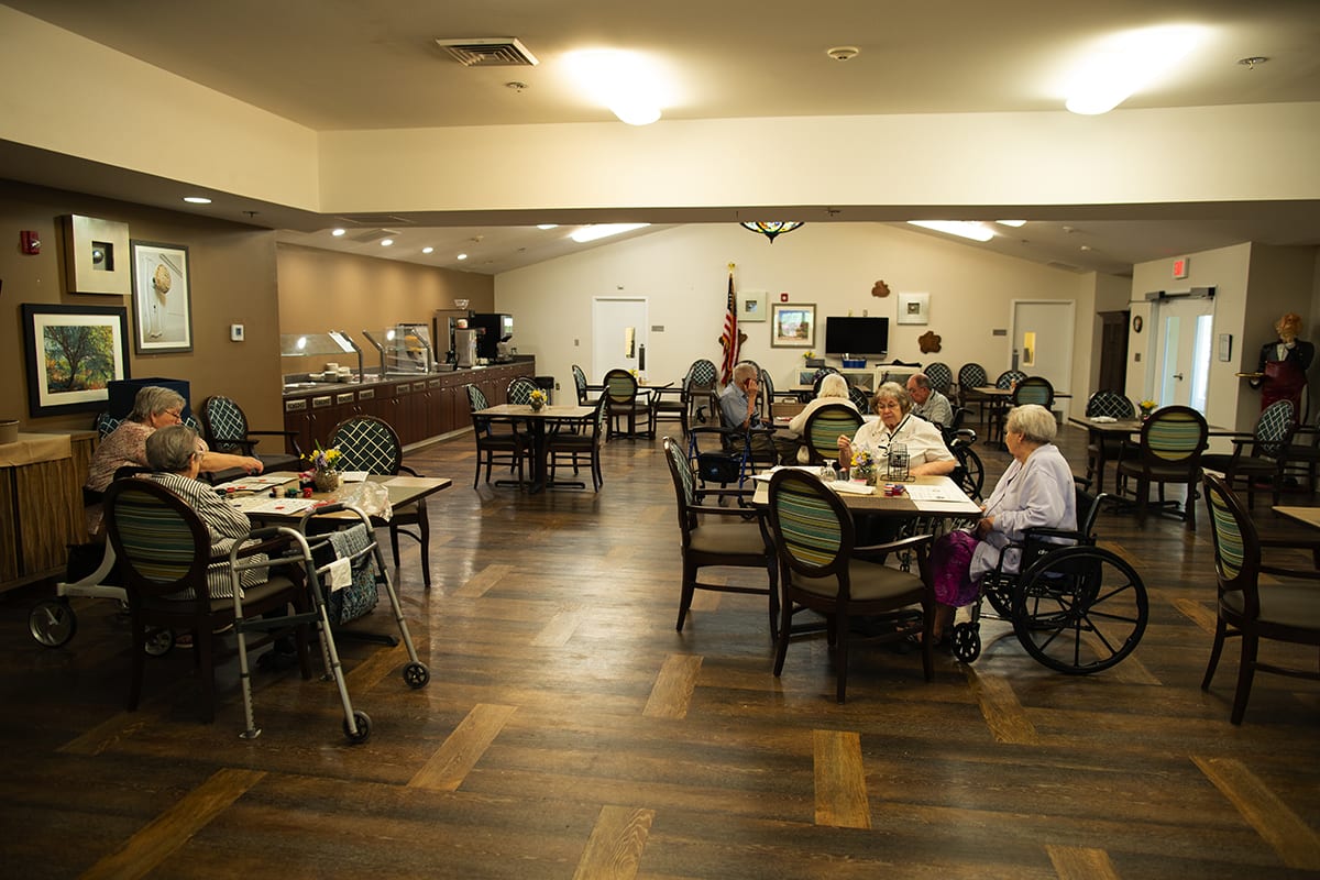 A dining hall with seniors seated at tables, some in wheelchairs, enjoying a meal in a community setting.