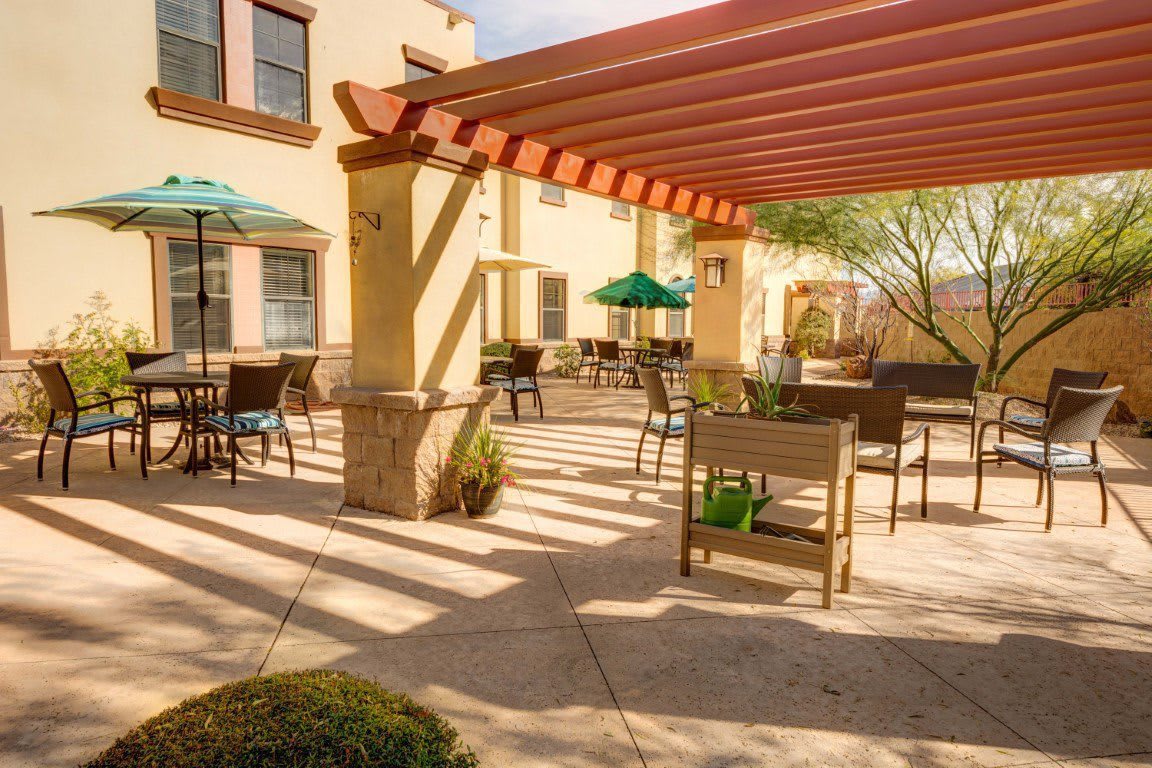 A sunny outdoor patio with a pergola, featuring tables and chairs near a building exterior.