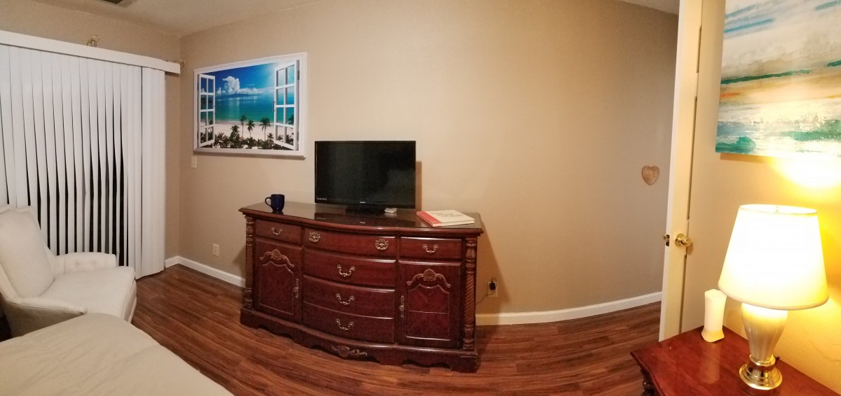 A cozy room featuring a wooden dresser with a television on top, accompanied by a cup and book, and a seaside painting hanging on the wall.