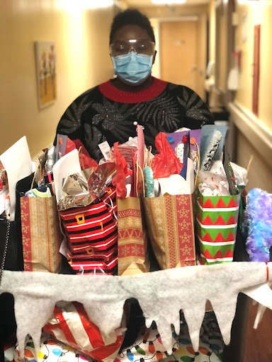 A masked individual stands in a hallway, proudly presenting a cart filled with beautifully wrapped holiday gifts.