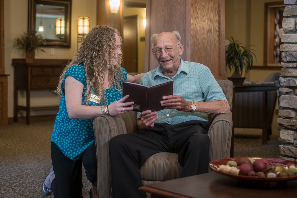 A caregiver assisting an elderly man in reading a book indoors.