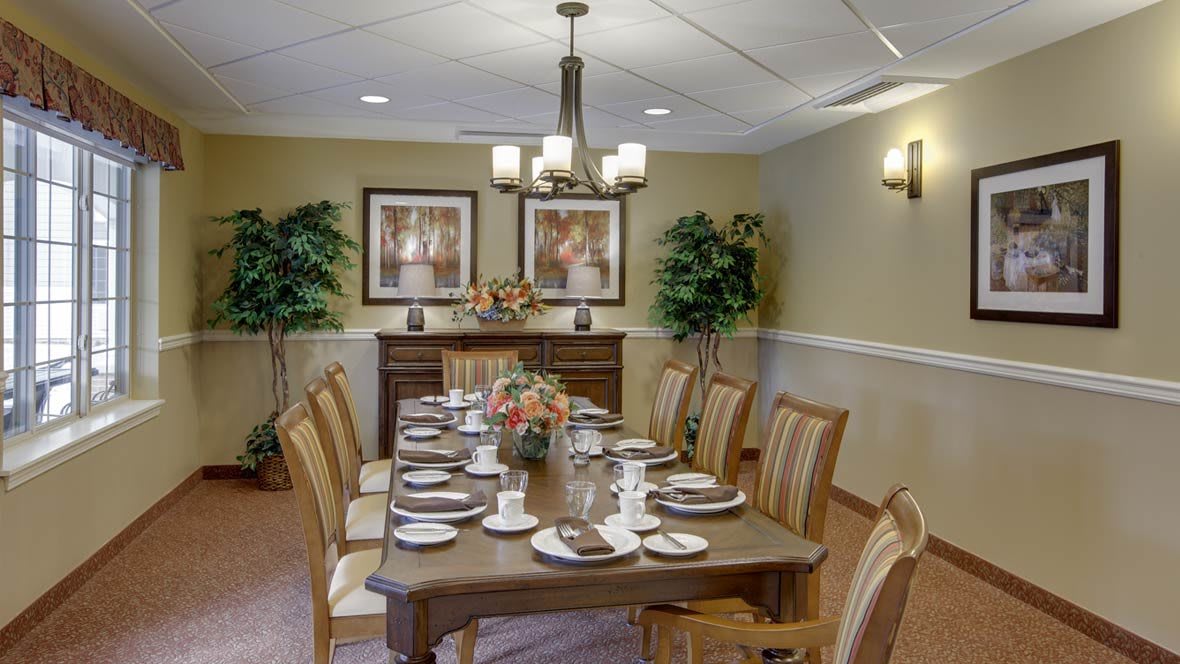 A formal dining room with a long table set for a meal, surrounded by chairs and decorated with plants and artwork.