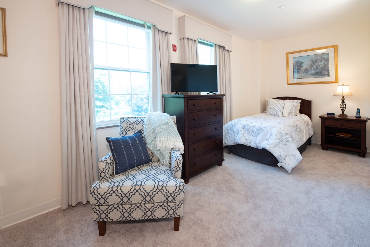 A cozy bedroom with a bed, chair, dresser, and a television near the window.