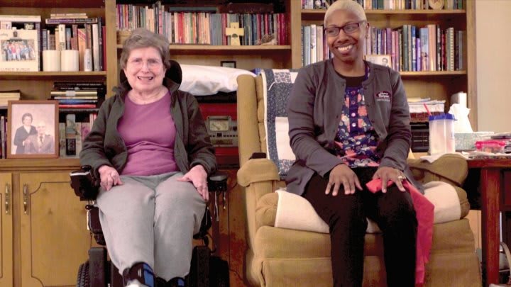 Two people smiling in a cozy living room with bookshelves in the background.