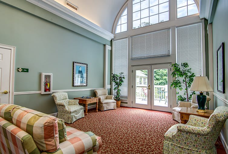 A cozy living room with large windows allowing natural light, furnished with comfortable seating and decorated with plants.