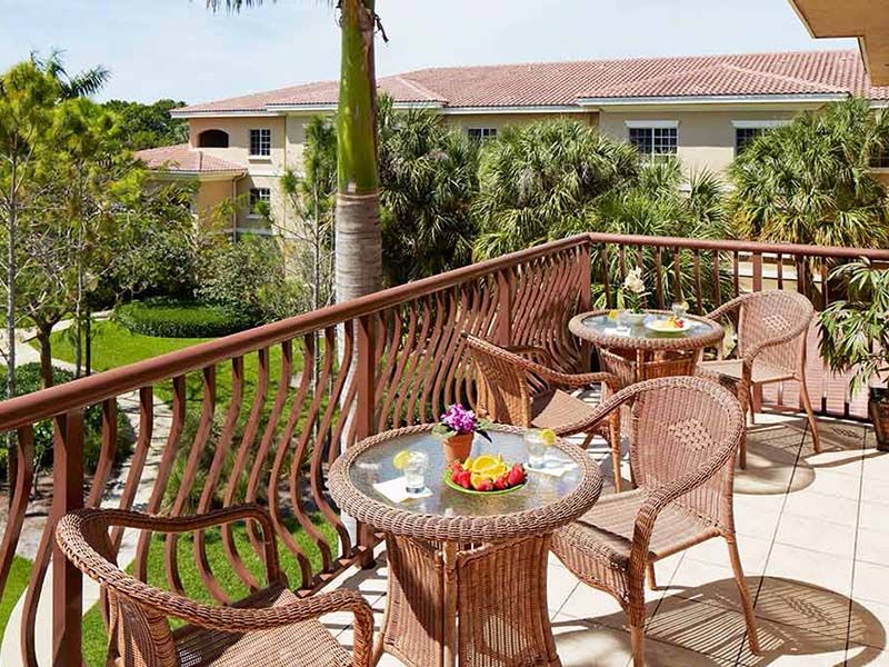 A sunny outdoor balcony with wicker tables and chairs overlooking a lush garden.