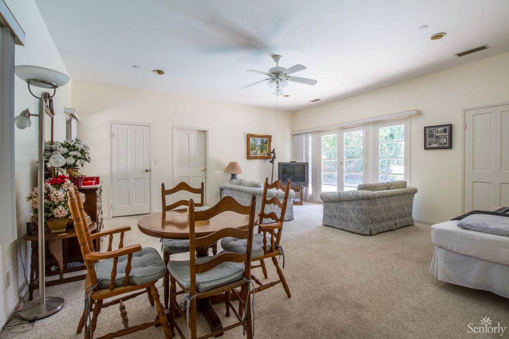A spacious living and dining area with a ceiling fan, carpet flooring, and large windows.