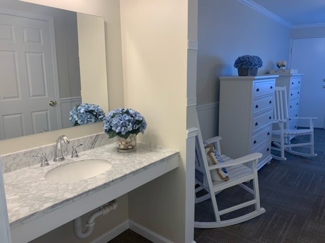 A cozy bathroom with a sink and vanity, complemented by a mirror and decorative flowers; a rocking chair and dresser are visible in the adjoining room.