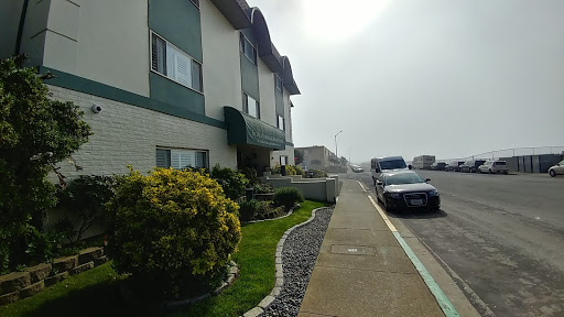 A view of a residential building along a foggy sidewalk with lush greenery and parked vehicles.