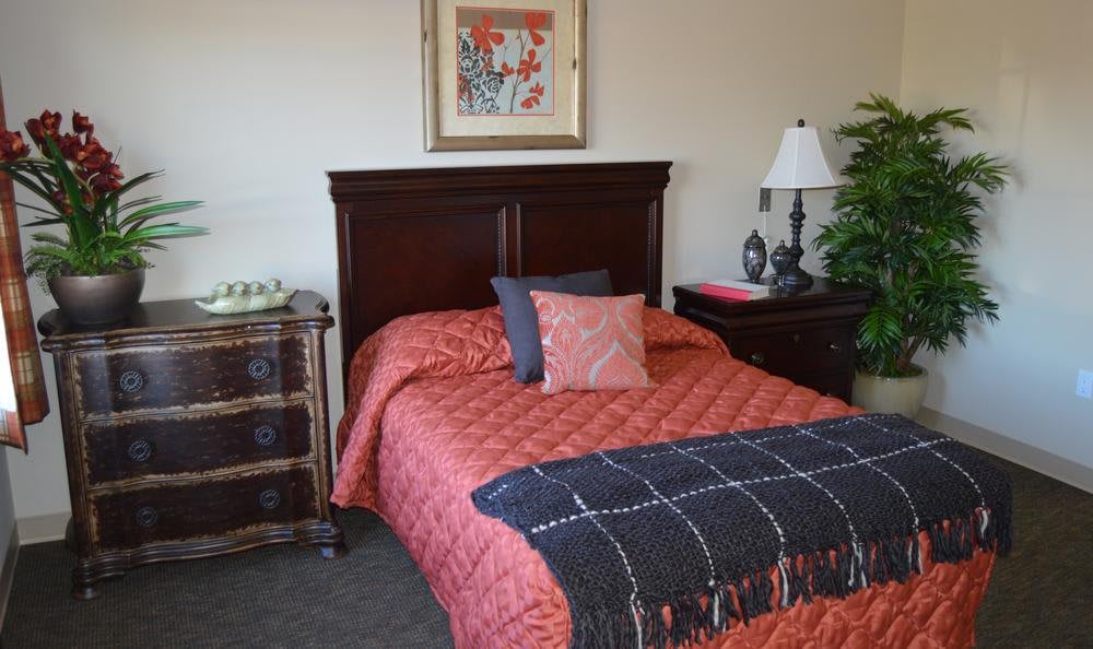 A neatly decorated bedroom with a red quilted bedspread and several pillows, featuring a wooden headboard, artwork on the wall, and a nightstand with a lamp.