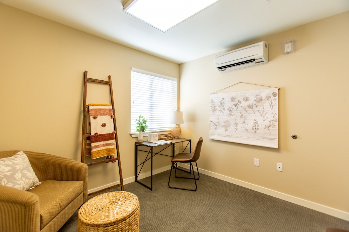 A cozy office space featuring a desk and chair, with natural light from a window, decorated with artwork and a ladder shelf.