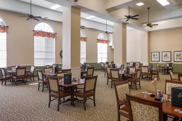 A spacious dining room with multiple tables and chairs, featuring large windows and overhead lighting.