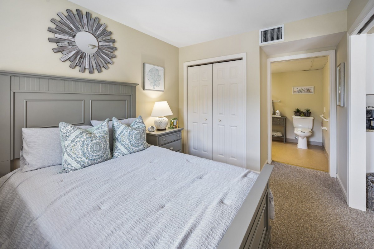 A cozy bedroom featuring neutral tones with a neatly made bed, a nightstand with a lamp, and a closet. The decor includes patterned pillows and a decorative mirror on the wall.