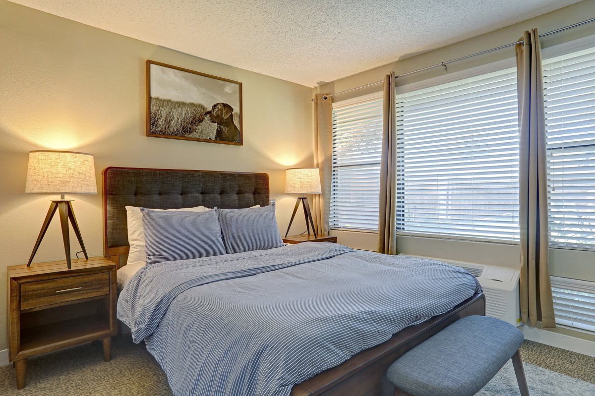 A cozy, well-lit bedroom featuring a neatly made bed with a tufted headboard, bedside tables with lamps, and large windows with blinds.