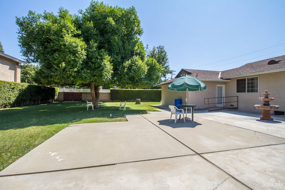 A sunny backyard with a patio featuring chairs and an umbrella, surrounded by grass and a large tree.