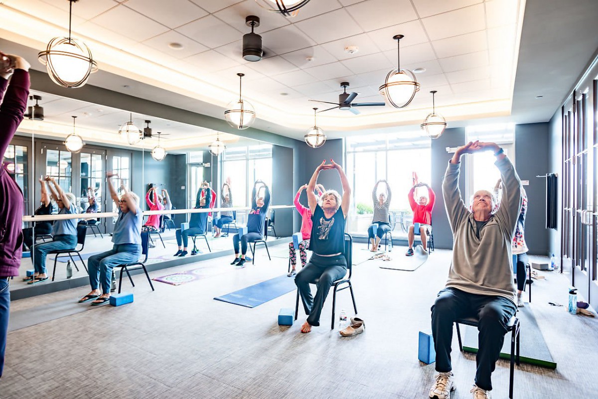 A group of seniors participates in a chair yoga class, performing stretching exercises indoors.