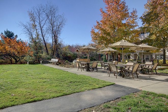 A scenic outdoor patio area with tables, chairs, and umbrellas surrounded by autumn trees and a well-maintained lawn.