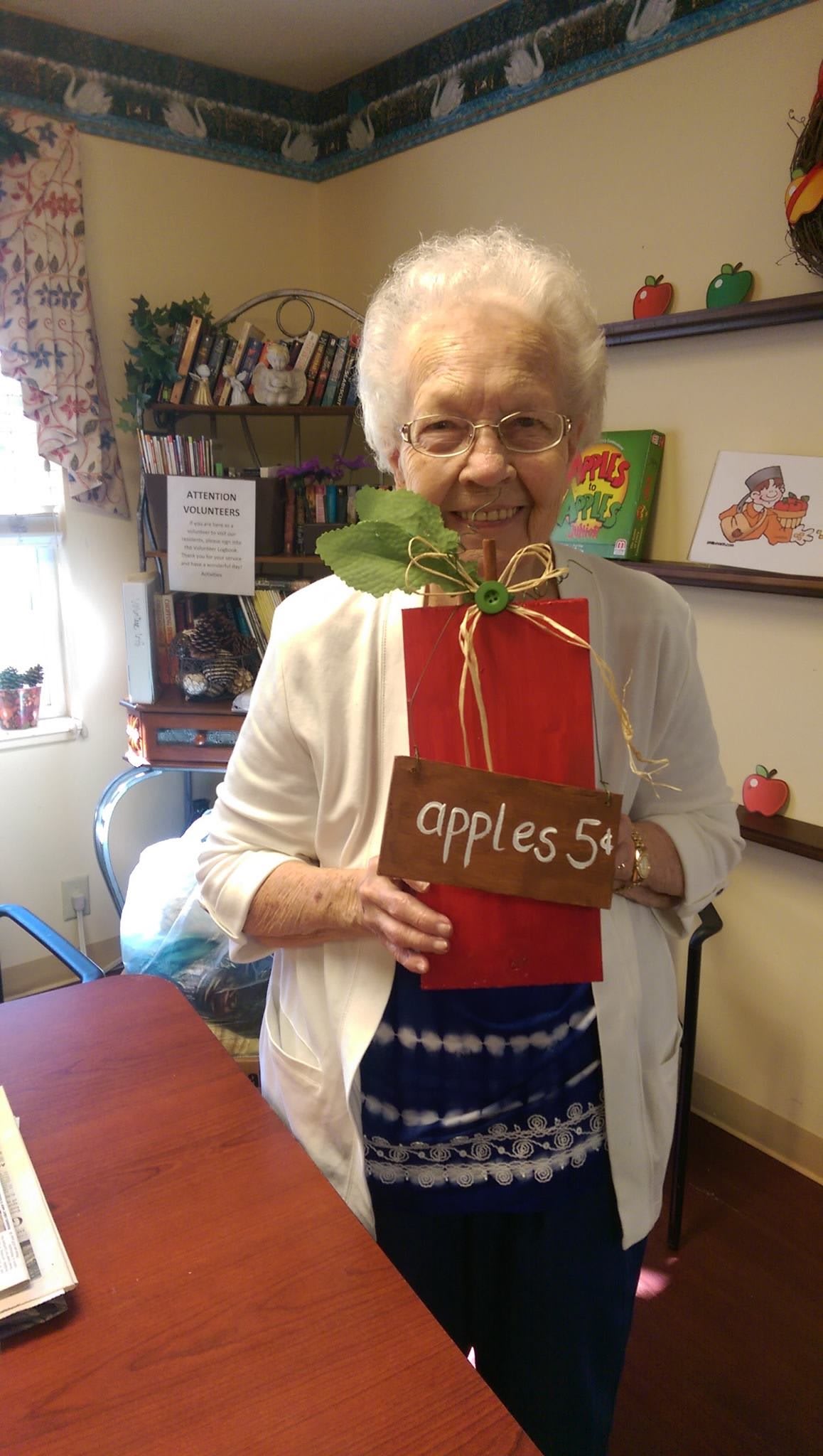 An elderly woman smiles while holding a gift wrapped in red packaging with a sign that says 'apples 5'.