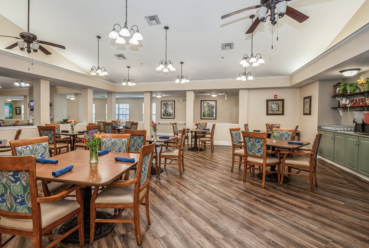 A well-lit dining room with multiple tables and chairs, featuring ceiling fans and chandeliers, and a wood floor.