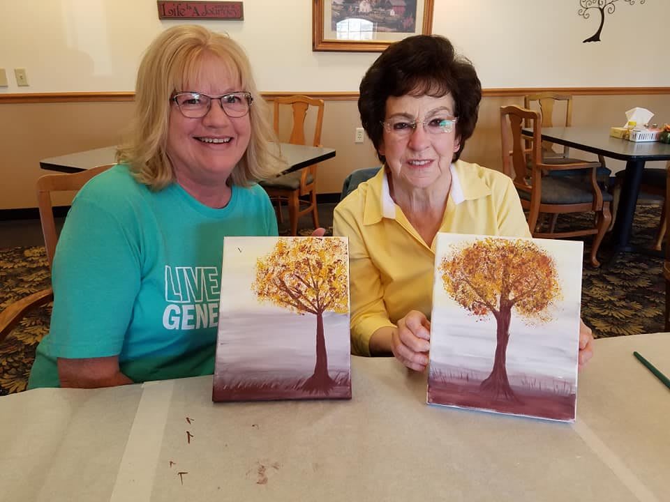 Two women proudly display their autumn tree paintings at an art workshop.