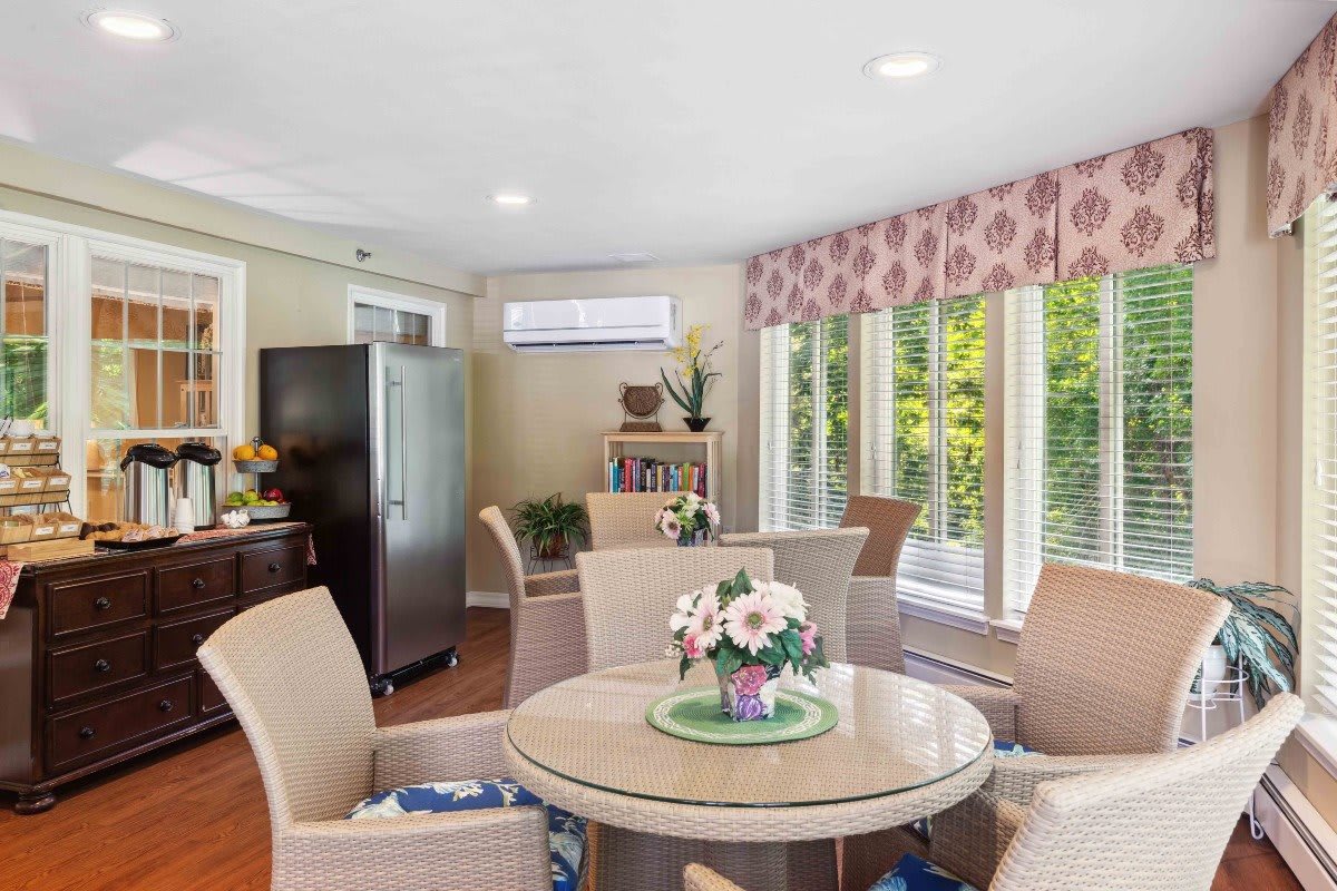 A cozy dining area with a round table and chairs, decorated with flowers and natural light streaming through large windows.