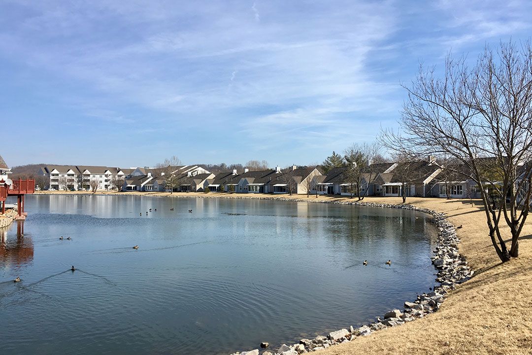 A peaceful lakeside scene with houses lining the shore under a partly cloudy sky.