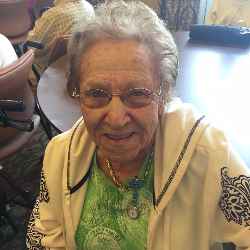 An elderly woman smiling indoors, wearing glasses and a casual outfit.