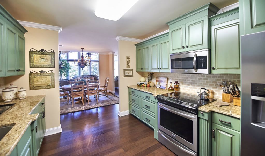 A modern kitchen with green cabinetry opens to an inviting dining area, showcasing hardwood flooring and sleek appliances.