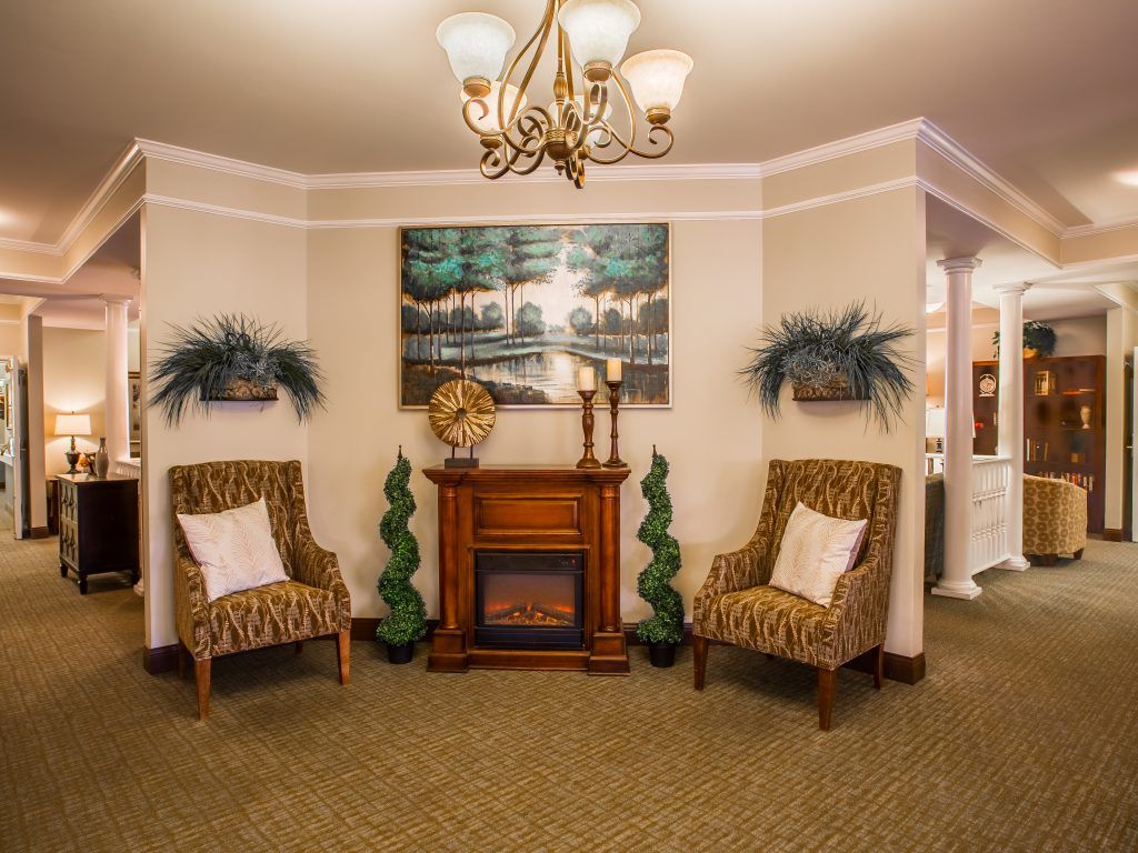 A cozy living room with a fireplace, two patterned chairs, artwork above the mantel, and decorative plants.