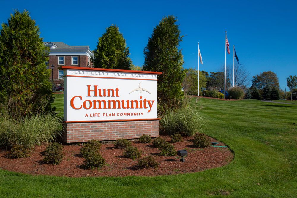 Entrance sign for Hunt Community, a senior living facility, with landscaped surroundings and flags in the background.