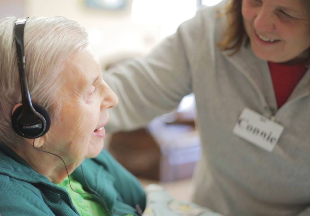 A caregiver interacts with an elderly person wearing headphones, providing support and companionship.
