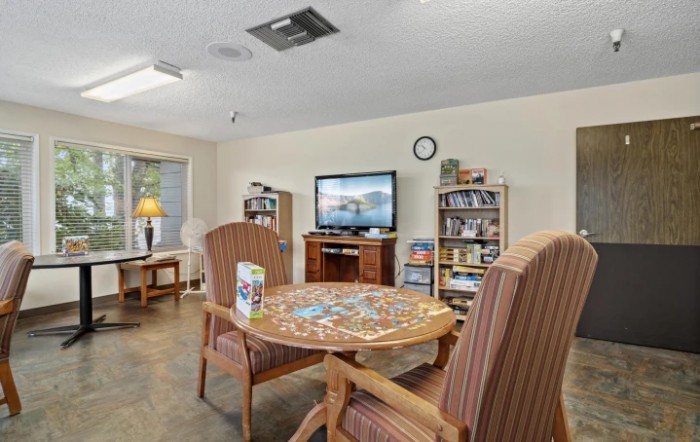 A cozy living room features a table with a puzzle, comfortable chairs, bookshelves, and a television.