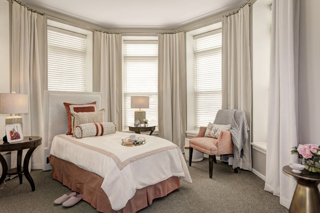 A cozy bedroom with a neatly made single bed, elegant curtains, and a stylish armchair beside a small lamp table.