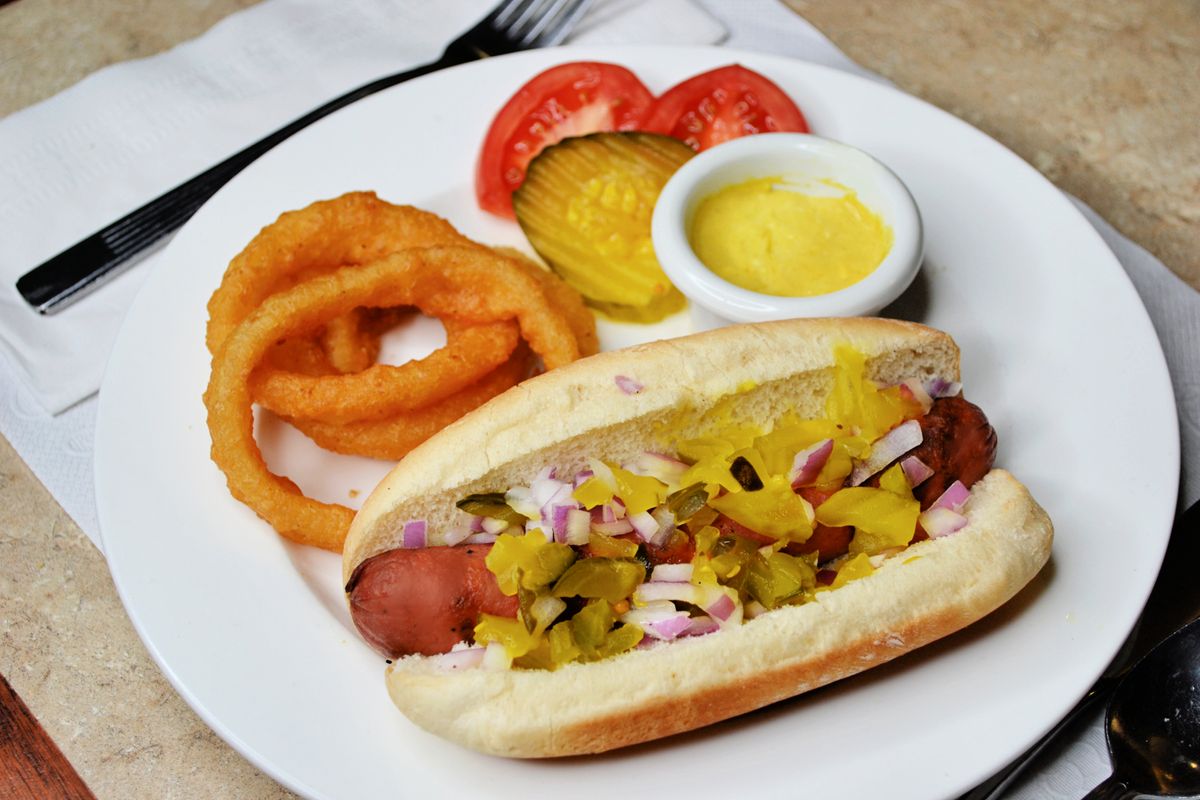 A hot dog topped with relish and onions, served with onion rings, pickles, tomatoes, and mustard.