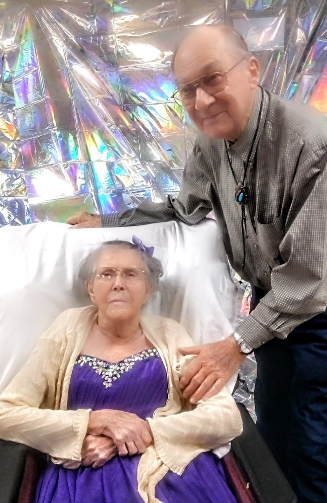 An elderly woman sits in a chair with a man standing beside her, surrounded by festive decorations.